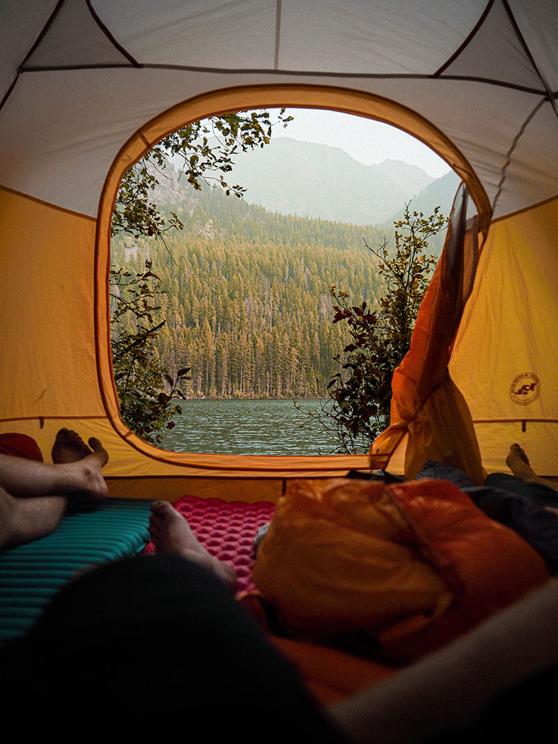 people lay in a tent looking out toward water and forested mountains