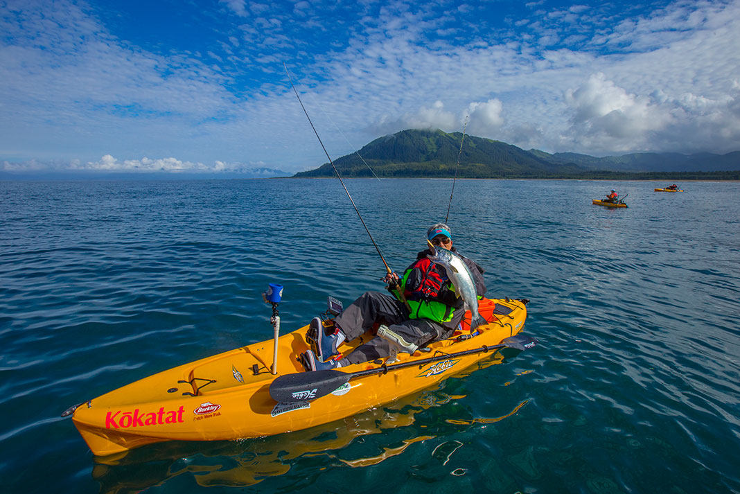 man in a yellow fishing kayak holds up a coho, or silver salmon, in front of the green, hilly Pacific coast with two other kayaks in the background