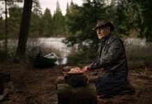 Algonquin fishing guide Frank Kuiack kneels and cleans fish at a campsite with canoe in background