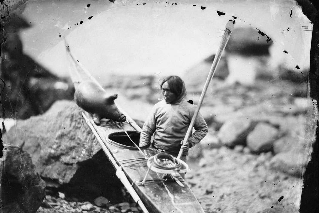 an unidentified Inuit man stands beside his kayak with seal hunting gear and a Greenland-style paddle
