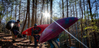 a group of whitewater kayakers walk through the forest carrying their boats