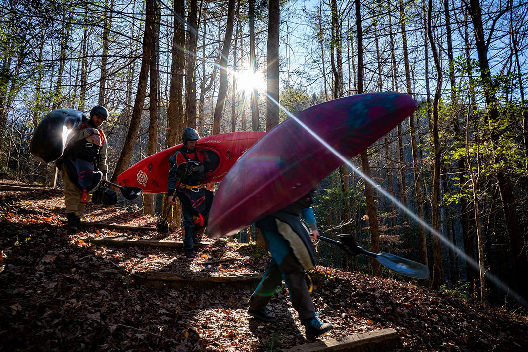 a group of whitewater kayakers walk through the forest carrying their boats