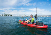 a young man paddles a red Perception Pescador 10.0 fishing kayak off a beach with palm trees