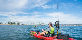 a young man paddles a red Perception Pescador 10.0 fishing kayak off a beach with palm trees