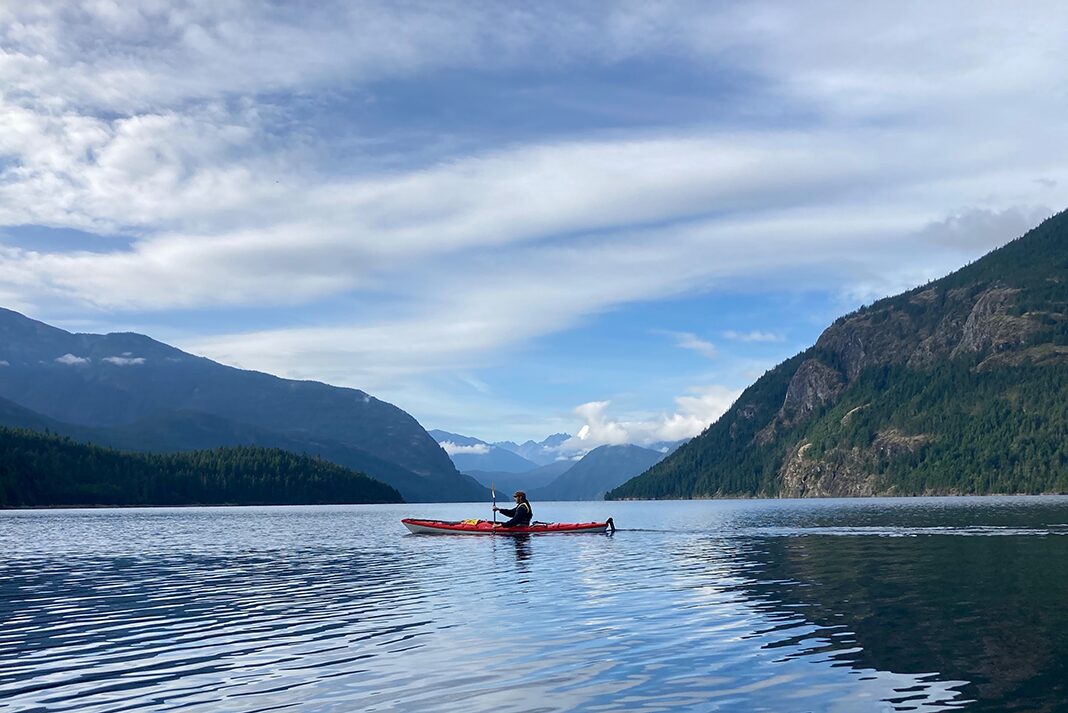 man paddles across calm mountain lake in kayak equipped with rudder