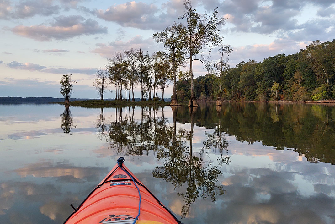 Bow of a kayak on mirror-like lake with trees in background