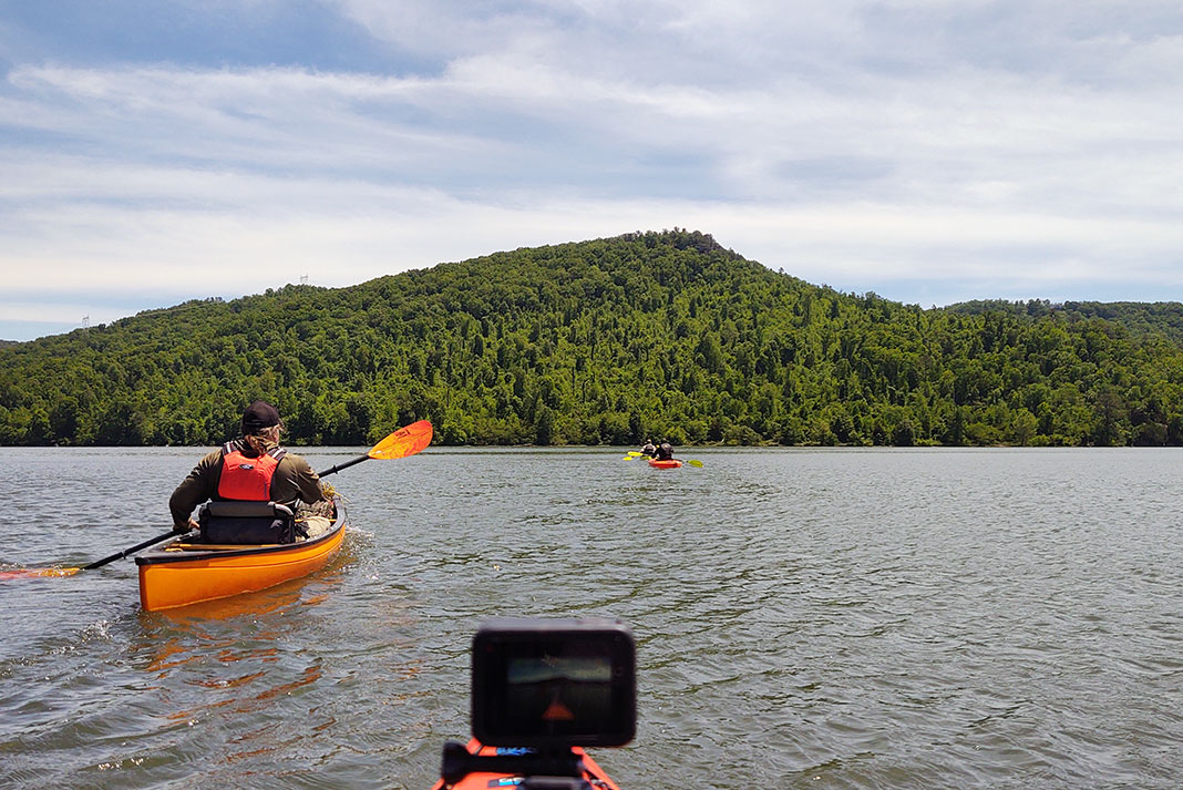 Three people paddling kayaks and canoes on a lake with a tree-covered hill in the background.