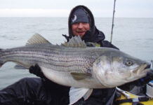 man holds up large striped bass caught while winter fishing as he tries to stay warm