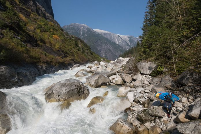Person carrying whitewater kayak around river full of boulders
