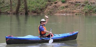 Man kneeling in solo canoe and waving at camera.