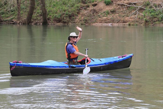 Man kneeling in solo canoe and waving at camera.