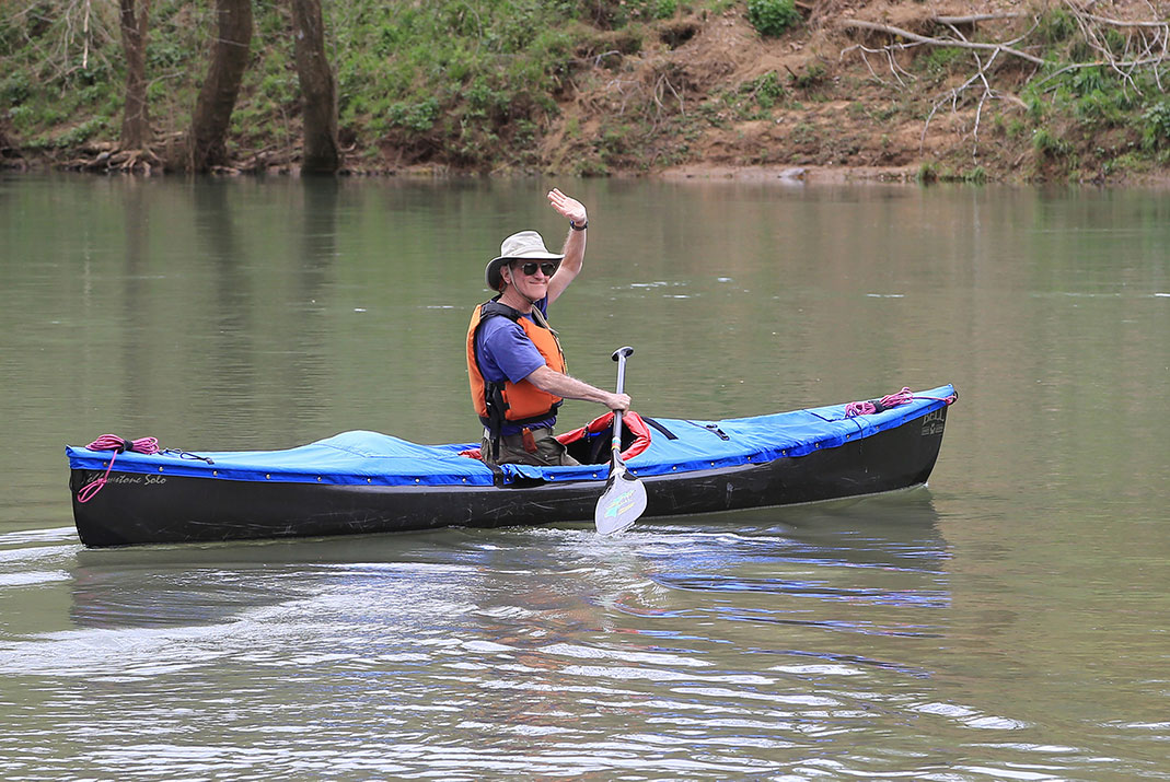 Man kneeling in solo canoe and waving at camera.