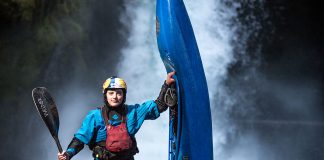 Woman wearing PFD, helmet, sprayskirt and drysuit. She is holding a kayak paddle in one hand, and a whitewater kayak in the other. She is standing in front of a waterfall.
