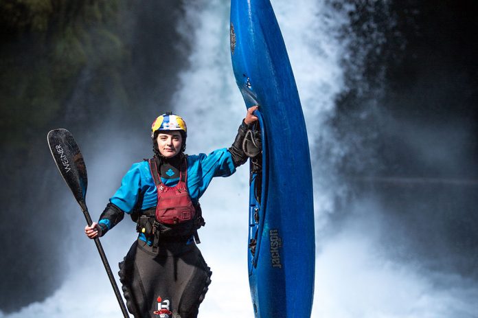 Woman wearing PFD, helmet, sprayskirt and drysuit. She is holding a kayak paddle in one hand, and a whitewater kayak in the other. She is standing in front of a waterfall.