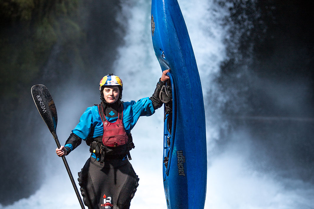 Woman wearing PFD, helmet, sprayskirt and drysuit. She is holding a kayak paddle in one hand, and a whitewater kayak in the other. She is standing in front of a waterfall.