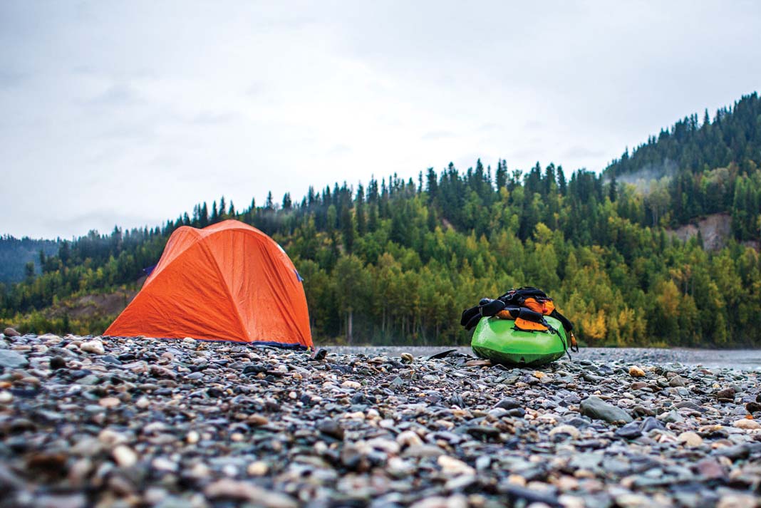 kayak and gear sit on pebbly riverside beach beside an orange tent