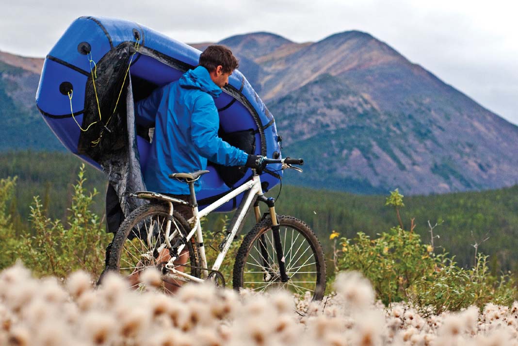 man carries a packraft on his shoulder while walking mountain bike through a field at foot of mountains