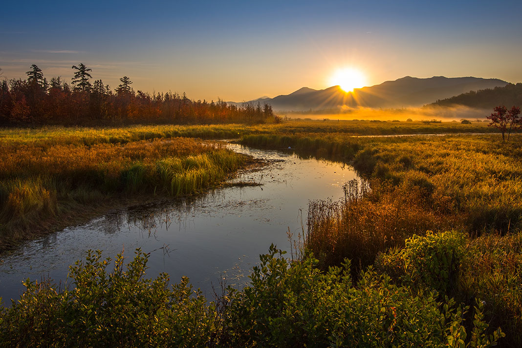 River running through marshland with mountain and sunset in background.