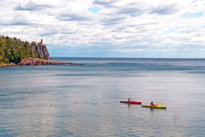 Two sea kayakers and lighthouse perched on rocky cliff in background.