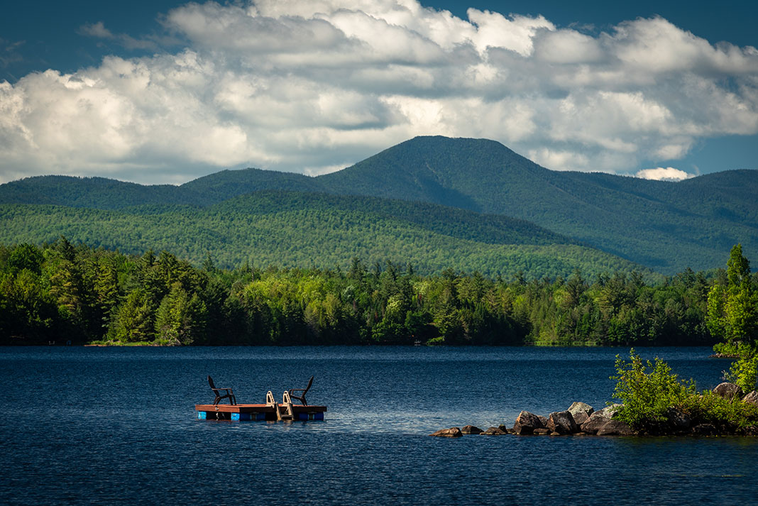Adirondack chairs on a floating raft on a lake with tree-covered hills in background.