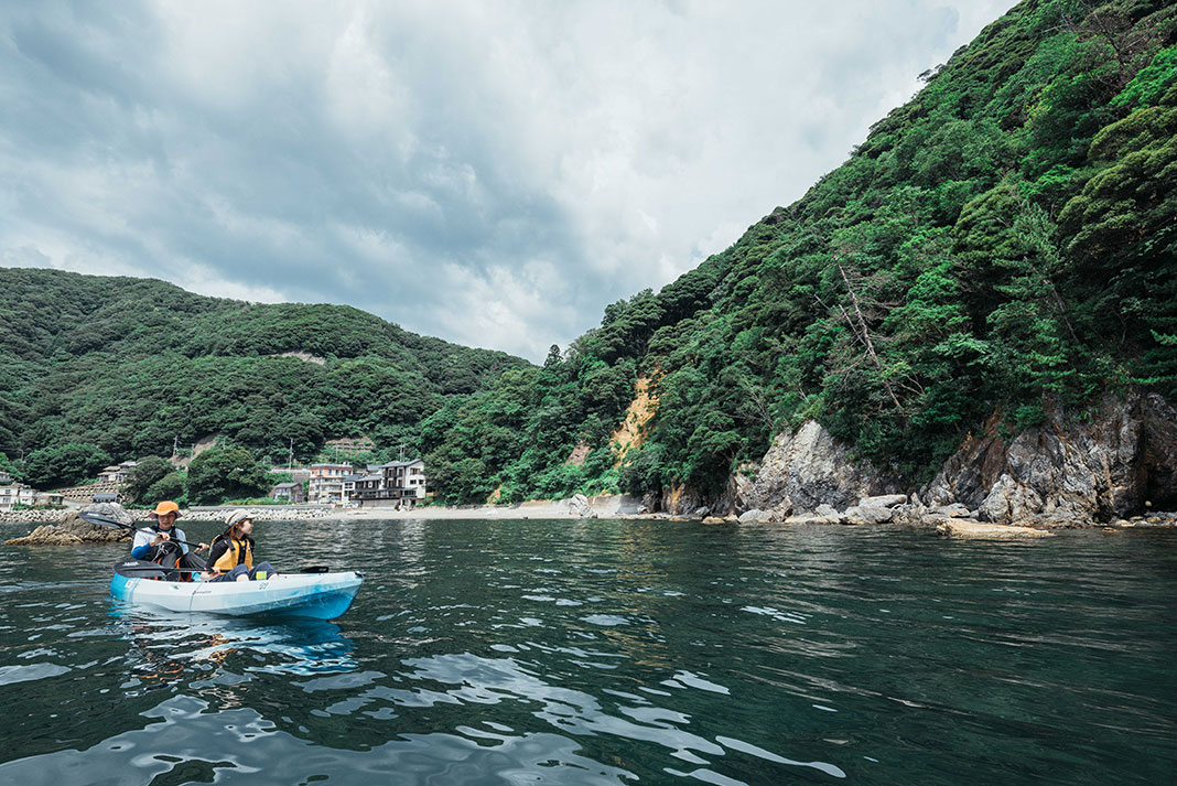 man and woman paddle a kayak on calm waters past lush, green hills in Japan