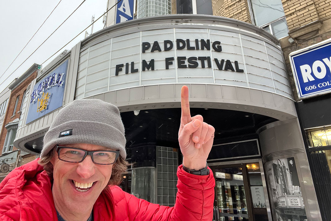 Man wearing winter hat and pointing at Paddling Film Festival sign on theater.