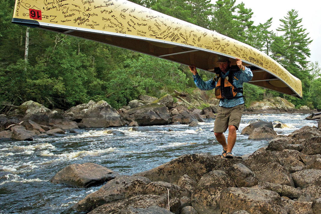 man walks along rushing river, carrying a canoe covered in signatures as part of a campaign to save the Boundary Waters
