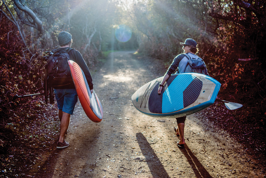 two men walk down a sunlit path carrying inflatable paddleboards and paddles