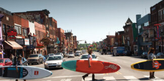 Three people crossing road with paddleboards under their arm.