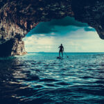 How To Choose The Right Inflatable PFD For Paddleboarding person paddleboarding into a watery cave entrance while wearing an inflatable PFD