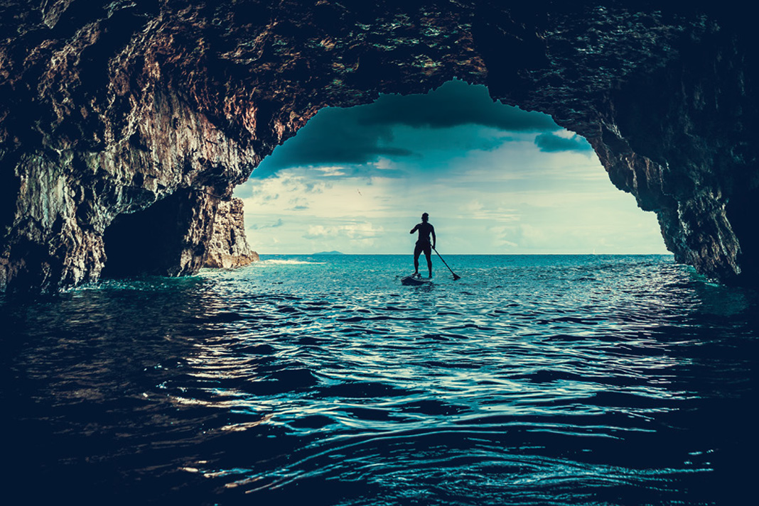 a person standup paddleboarding near a cave entrance while wearing an inflatable PFD