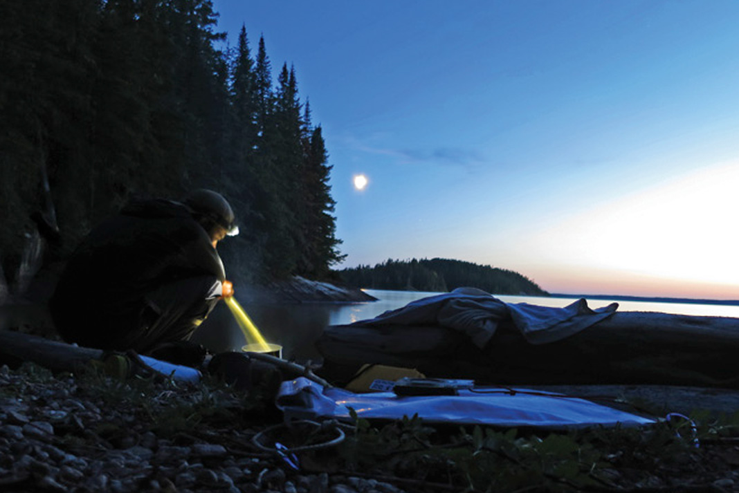 man cooks by headlamp at a riverside camp site near midnight