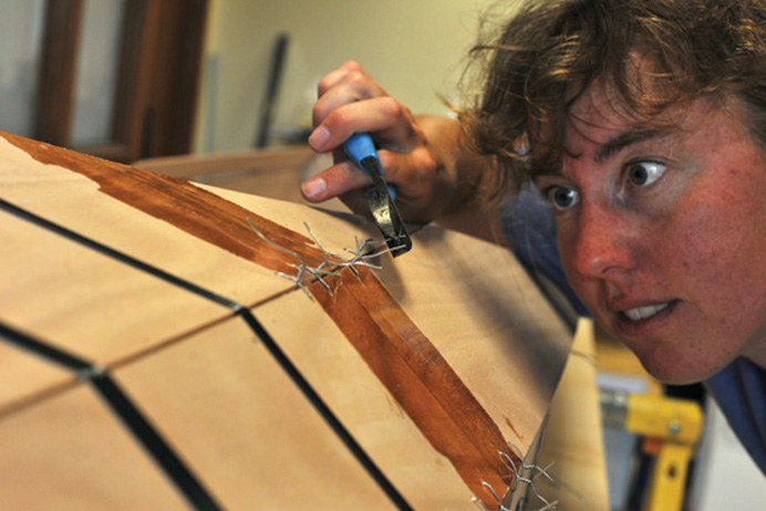 woman stares at wooden kayak as she works on building it with pliers and metal wire