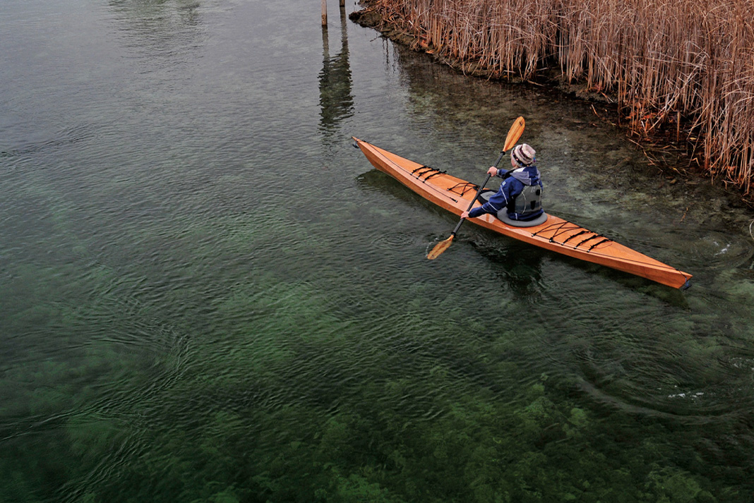 person paddles a wooden kayak from Pygmy Boats in shallow water with autumn reeds behind her