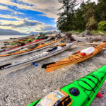 a group of touring kayaks rest on a pebble beach under a vibrant sky