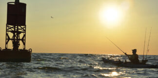 kayak angler is silhouetted against the seaside afternoon sun as he casts a lure near a buoy