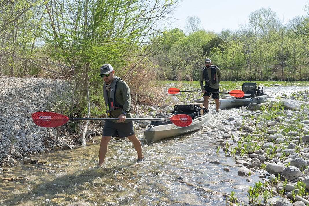 two men drag their fishing kayaks down the shallow, rocky Frio River