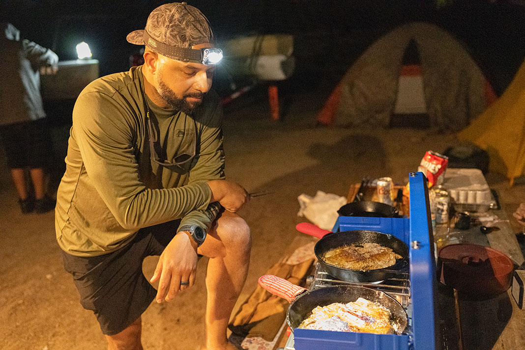 man wearing headlamp stands over a blue cook stove with food cooking at night
