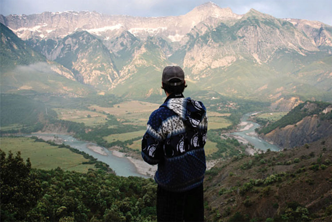 a man in hat and sweater looks over a river valley surrounded by mountains from a high vantage point