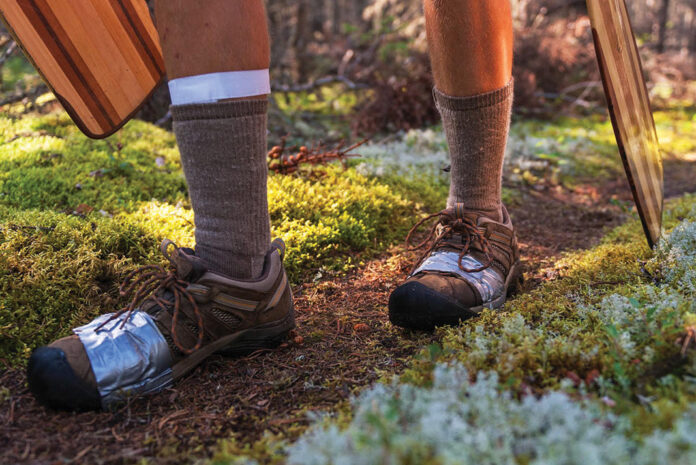 person carrying paddles along a mossy portage trail with shoes wrapped in duct tape