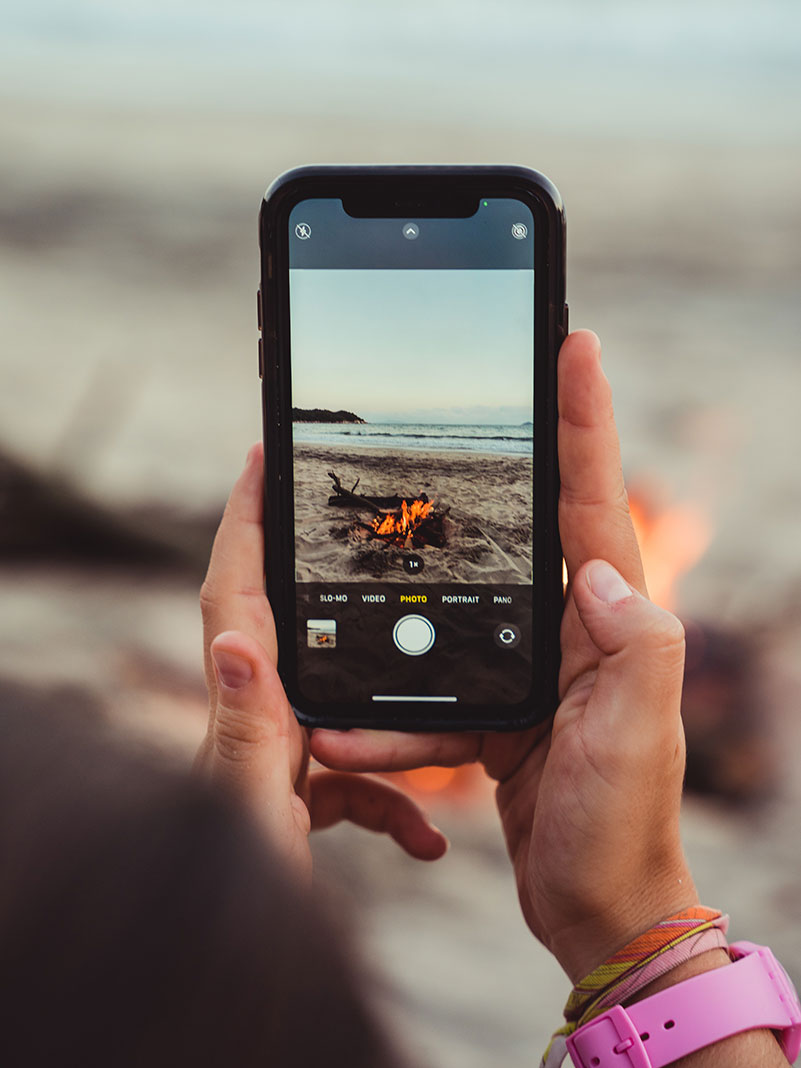 person uses a cell phone to take a photo of a beach campfire in the wilderness