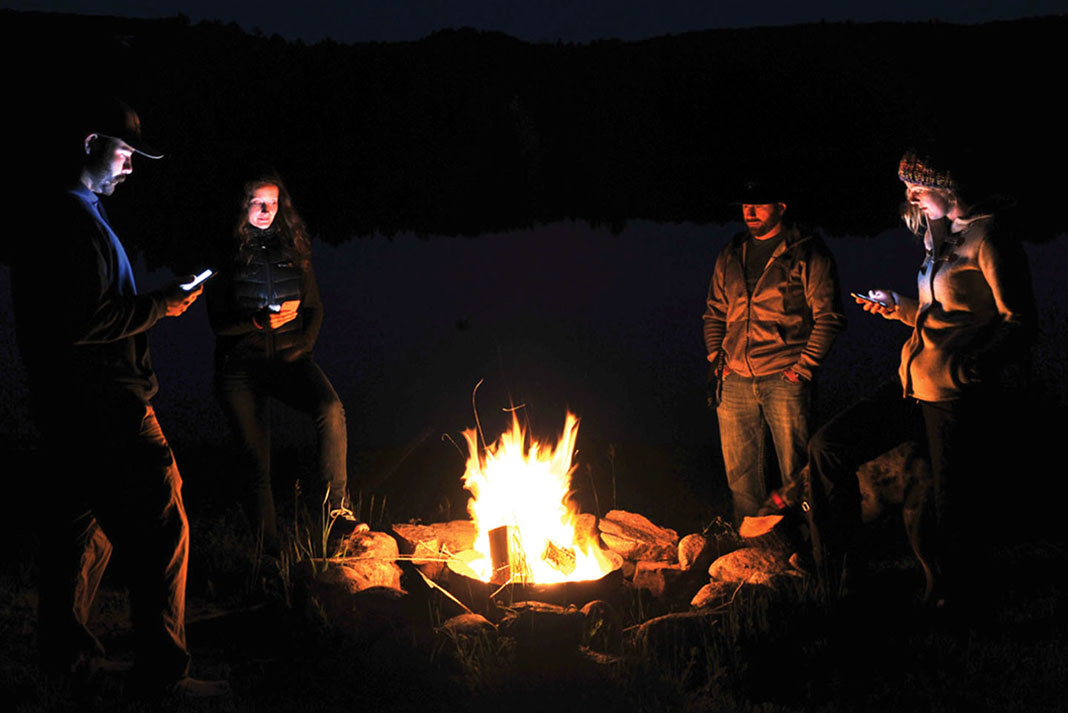 people stand around a campfire in the wilderness while using their cell phones