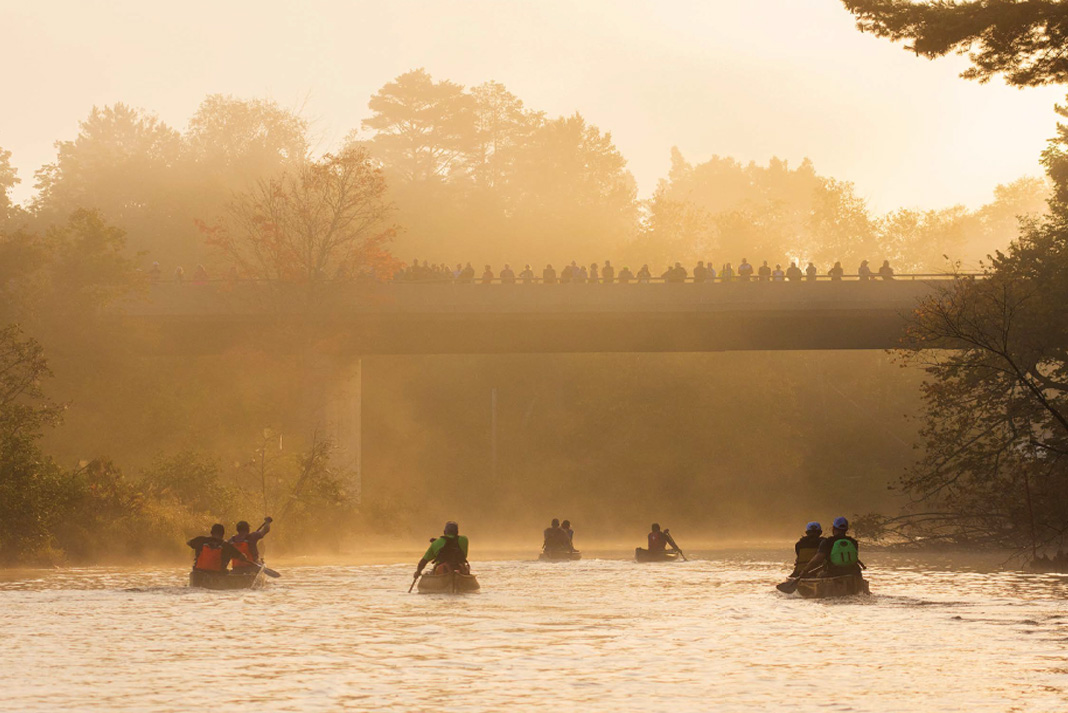 a group of racing canoeists paddle in the mist with spectators watching from a bridge overhead