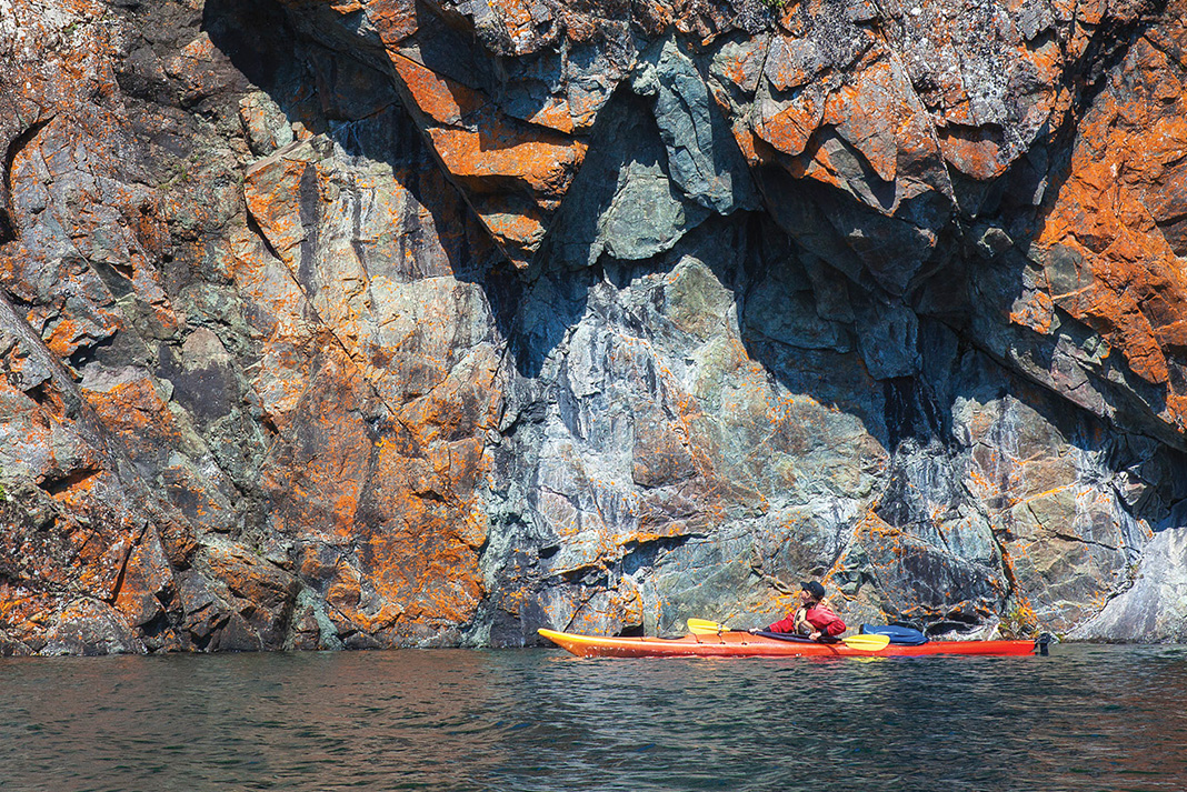 person sea kayaking along dramatic orange and grey rocky shoreline