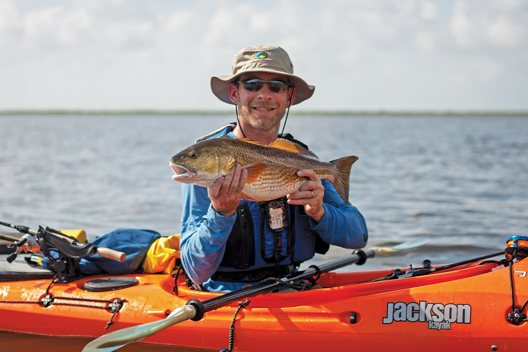 man holds up a fish caught from an orange Jackson Kayak