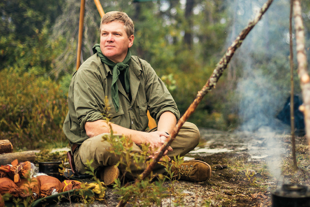 Ray Mears sits cross-legged in front of a smoking cook pot at a campsite in Wabakimi Provincial Park