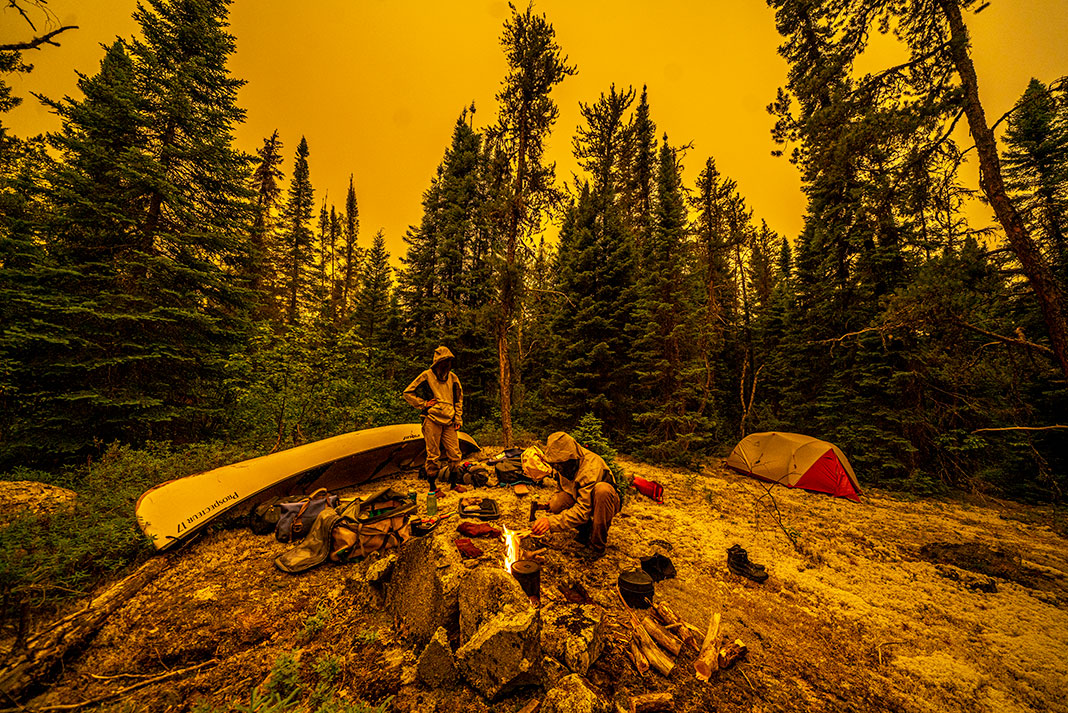two people in bug jackets at a campsite along an ancient canoe to Hudson Bay with everything tinted yellow by the smoke of a nearby forest fire