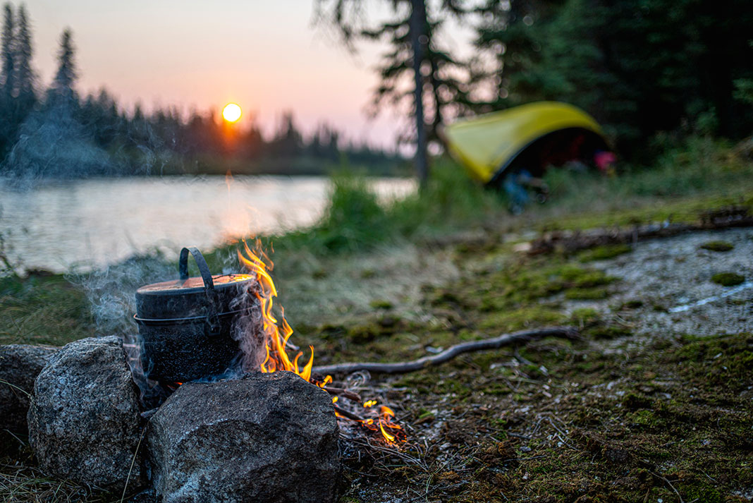 a covered pot sits on a campfire at dusk with beached yellow canoe and river behind