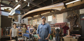 nick offerman stands in his woodshop surrounded by staff with hand-crafted canoe hanging overhead