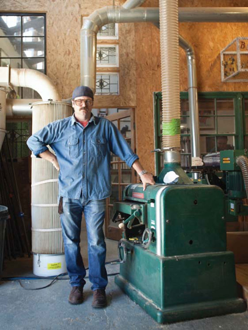 Nick Offerman wears hat and goggles while standing beside machinery in his woodshop